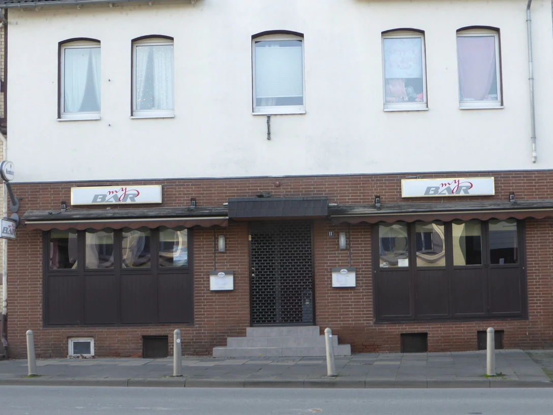 Fassade einer Stadtbar mit dunklen Fenstern, Markise und zentralem Eingang an der Straße.