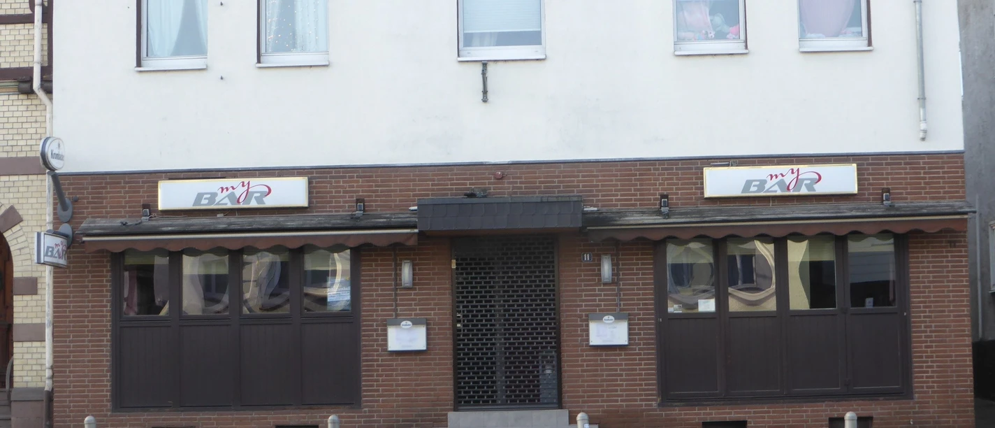 Fassade einer Stadtbar mit dunklen Fenstern, Markise und zentralem Eingang an der Straße.