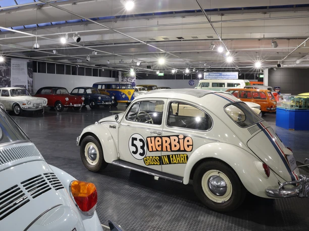Blick in die Ausstellung des Automuseum Volkswagen Innenraum mit alten VW-Autos. Vorn steht ein weißer Käfer mit Nummer 53.