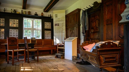 heidemuseum-wilsede.jpg Museumzimmer mit Holzmöbeln, Wiege, Tisch, Stühlen und Kachelwand in historischem Bauernhaus.
