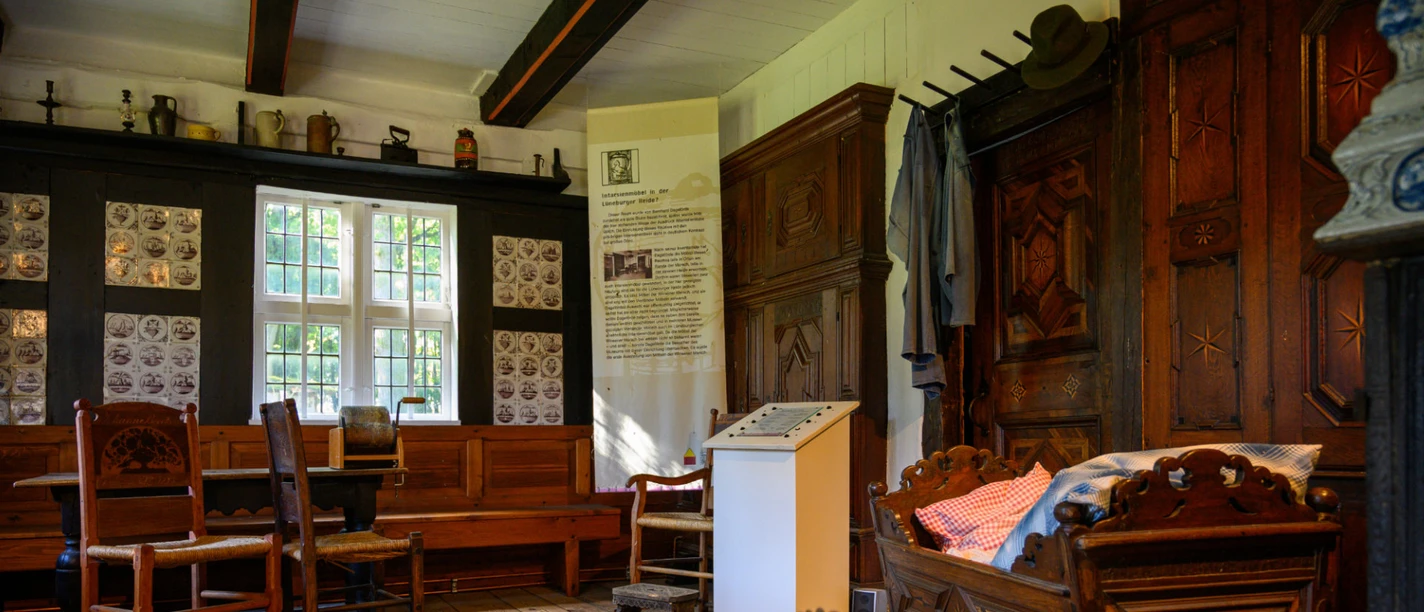 heidemuseum-wilsede.jpg Museumzimmer mit Holzmöbeln, Wiege, Tisch, Stühlen und Kachelwand in historischem Bauernhaus.
