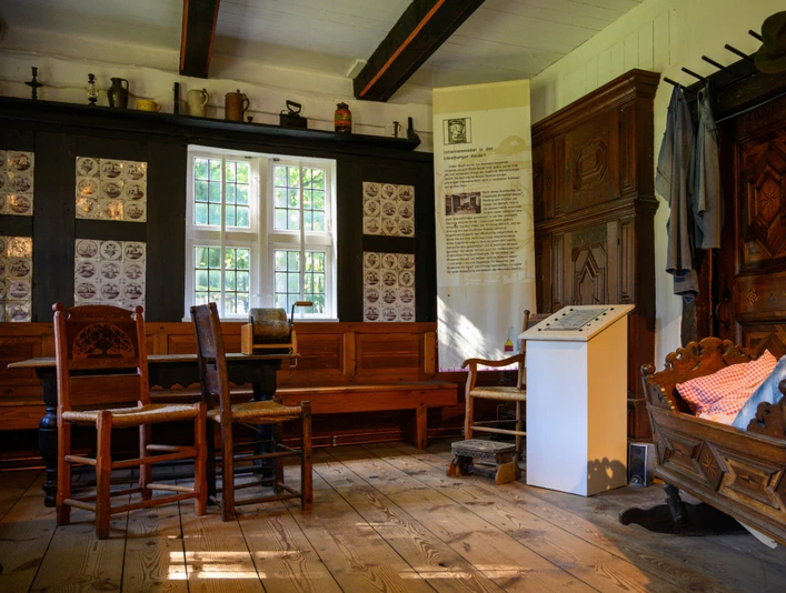 heidemuseum-wilsede.jpg Museumzimmer mit Holzmöbeln, Wiege, Tisch, Stühlen und Kachelwand in historischem Bauernhaus.