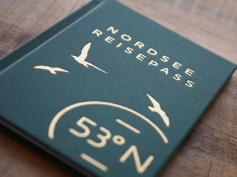 TANO-Nordsee-Reisepass.jpg Grünes Heft mit goldener Prägung NORDSEE REISEPASS und Möwenmotiv auf hölzerner Fläche.