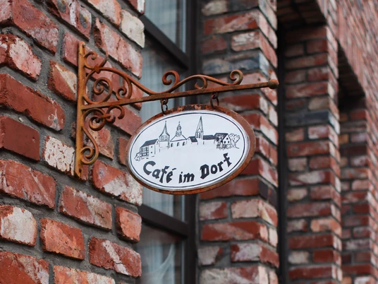 Schild "Café im Dorf" in Haan-Gruiten Schmiedeeisernes Ovalschild "Café im Dorf" mit Dorf-Silhouette an einer rotbraunen Ziegelwand.