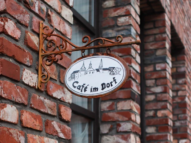 Schild "Café im Dorf" in Haan-Gruiten Schmiedeeisernes Ovalschild "Café im Dorf" mit Dorf-Silhouette an einer rotbraunen Ziegelwand.