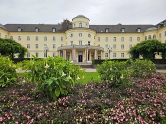 Gelbes Kurgebäude mit Rundbau hinter Blumenbeeten und grünen Sträuchern im Park