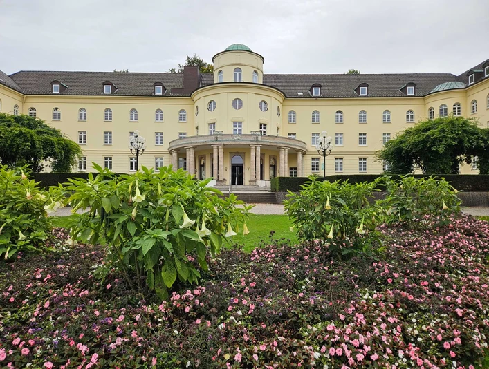 Bad Hermmansborn Gelbes Kurgebäude mit Rundbau hinter Blumenbeeten und grünen Sträuchern im Park