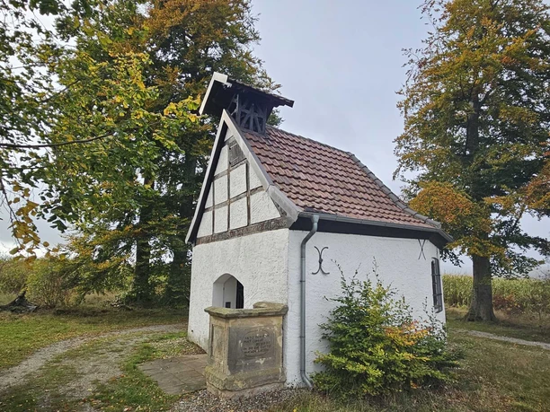 Kleine weiße Fachwerkkapelle mit Glockenstuhl, umgeben von herbstlichen Bäumen.