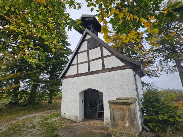 Kleine weiß verputzte Kapelle mit Fachwerk und Glockenstuhl zwischen großen Laubbäumen.