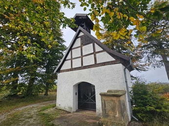 Dringenberg-Schonlaukapelle Kleine weiß verputzte Kapelle mit Fachwerk und Glockenstuhl zwischen großen Laubbäumen.