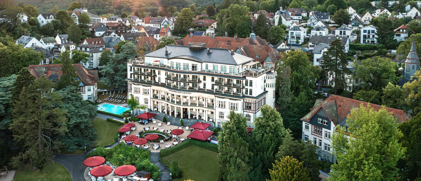 Falkenstein Grand Autograph Collection - Luftaufnahme Luftaufnahme des Falkenstein Grand Hotels, einem weißen Gebäude in einer grünen, hügeligen Landschaft. Rote Sonnenschirme beleben die Terrasse.