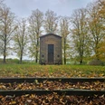 Herste-Rommenhöller-Ehrenmal- Steingebäude vor herbstlichen Bäumen, mit Laub bedeckten Stufen und zwei Bänken.