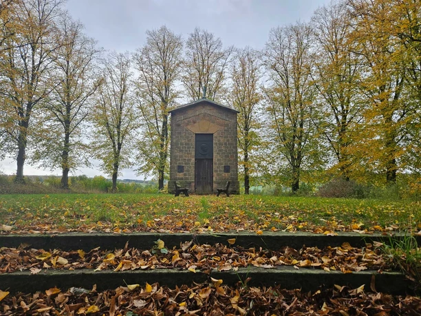 Herste-Rommenhöller-Ehrenmal Steingebäude vor herbstlichen Bäumen, mit Laub bedeckten Stufen und zwei Bänken.