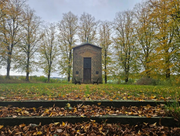Herste-Rommenhöller-Ehrenmal- Steingebäude vor herbstlichen Bäumen, mit Laub bedeckten Stufen und zwei Bänken.
