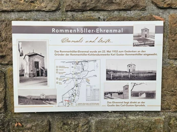 Infotafel zum Rommenhöller-Ehrenmal mit historischen Fotos und Lageplan vor Steinmauer.