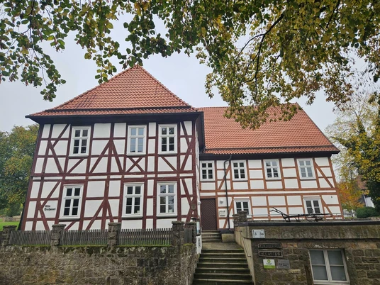 Fachwerkhaus mit rotem Dach und Treppe im Vordergrund, umgeben von Bäumen.