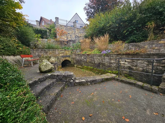 Steinmauern, Stufen und eine kleine Quellöffnung in einem bepflanzten Hang mit Bank.