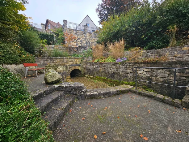 Steinmauern, Stufen und eine kleine Quellöffnung in einem bepflanzten Hang mit Bank.