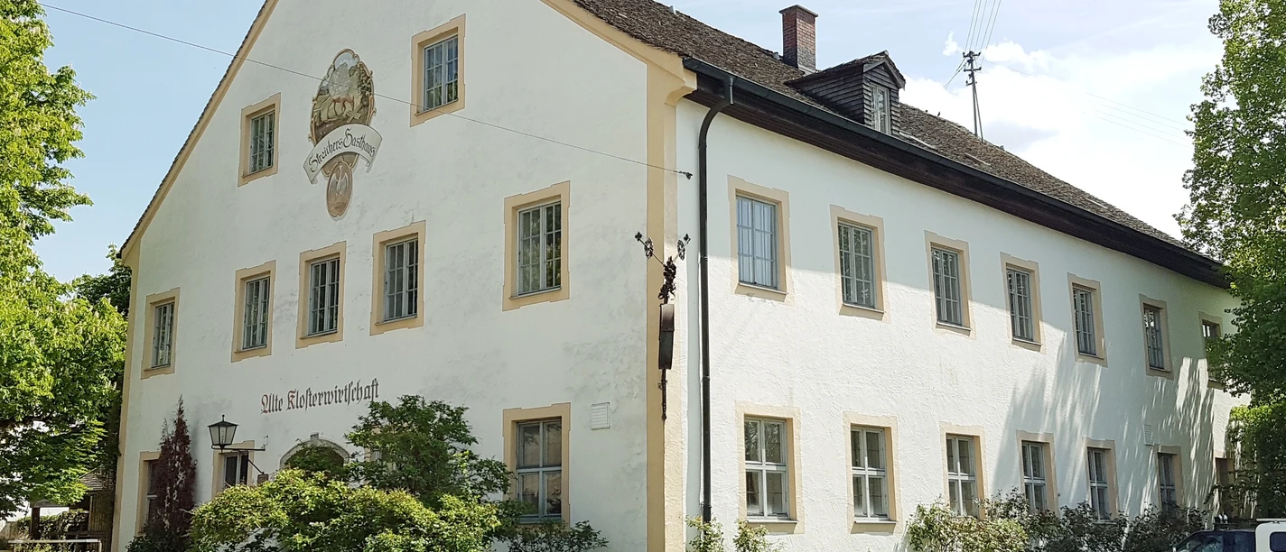 Klosterwirtschaft Polling Großes historisches Haus mit weißer Fassade. Heller Tag. Bäume umgeben das Gebäude.