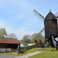 Mühlengarten_Salder_HD_16zu9.JPG Historische Bockwindmühle mit Holzbau, flankiert von Sitzbänken und Fachwerkhaus im Grünen.