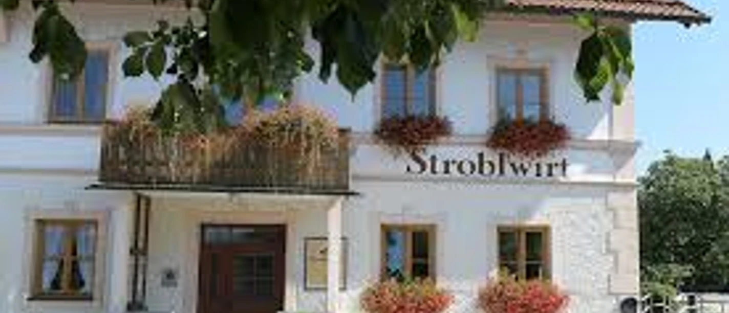 Stroblwirt Weißes Haus mit vielen Blumen und dem Schriftzug Stroblwirt unter grünen Blättern.