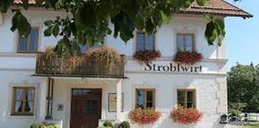 Stroblwirt Weißes Haus mit vielen Blumen und dem Schriftzug Stroblwirt unter grünen Blättern.