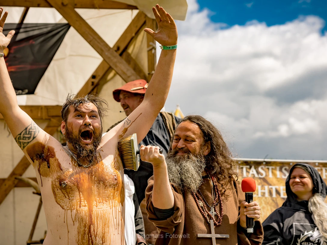 Markteröffnung Ein Mann wird bei einer Mittelaltershow lachend mit Teer eingerieben, umgeben von kostümierten Darstellern.