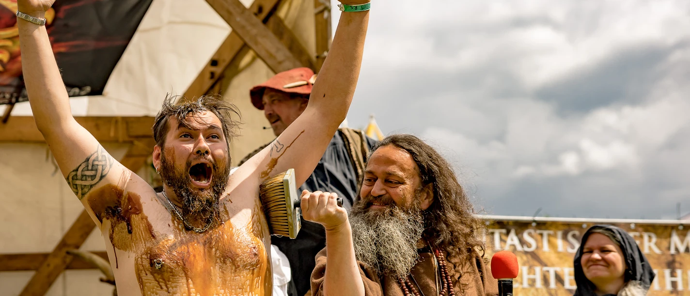 Markteröffnung Ein Mann wird bei einer Mittelaltershow lachend mit Teer eingerieben, umgeben von kostümierten Darstellern.