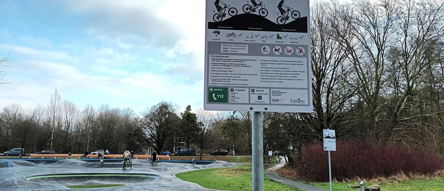Die Pumptrackanlage in Salzgitter-Gebhardshagen Infoschild und Holzbank an einer asphaltierten Pumptrackanlage mit Radfahrern.