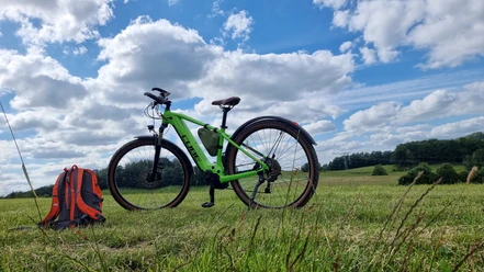 Grünes Fahrrad und roter Rucksack auf Wiese vor bewölktem Himmel.