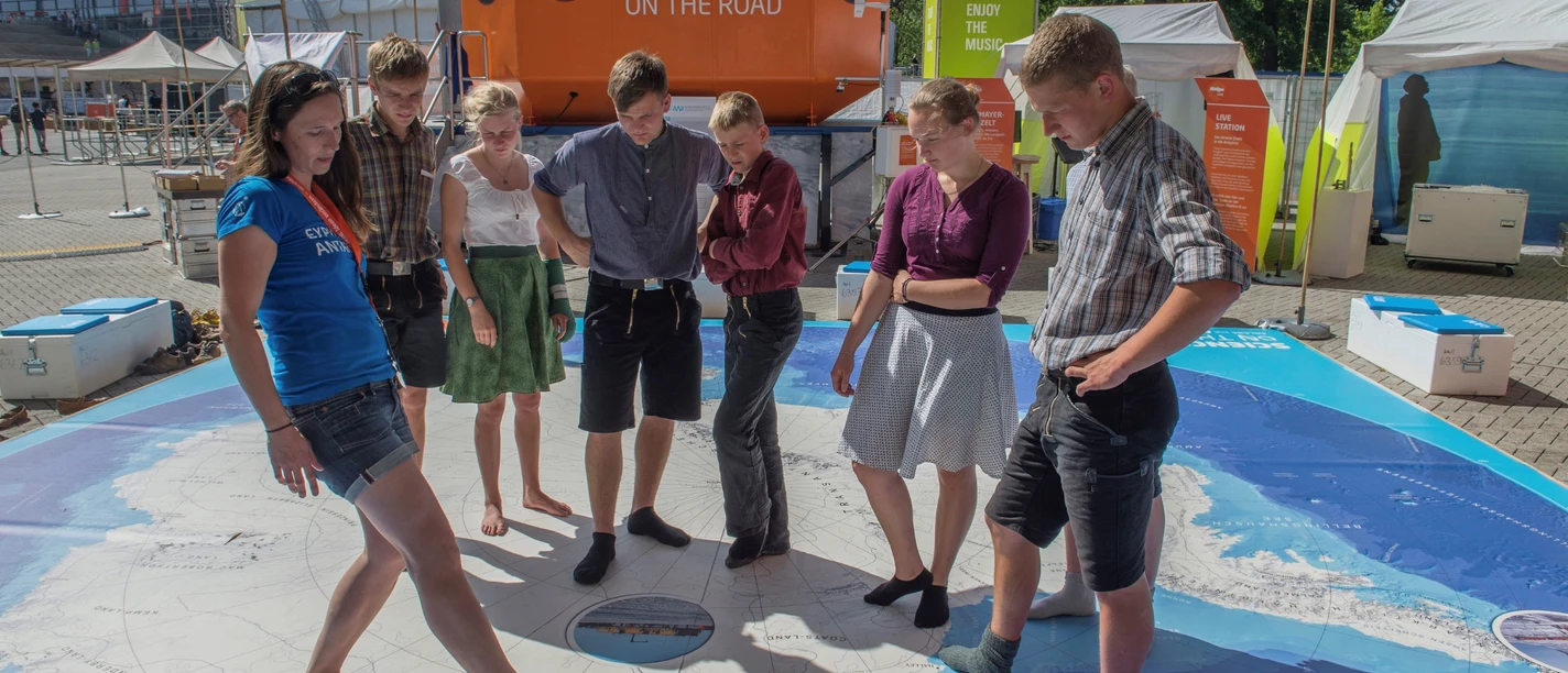 H. Grobe, Sience on the Road Gruppe junger Menschen erkundet eine große Weltkarte im Freien vor einem mobilen Forschungslabor.