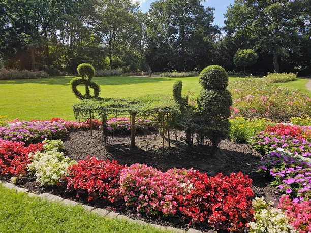 Grüne Formschnittfigur am Klavier aus Buchsbaum, umgeben von bunten Blumenbeeten im Bürgerpark.