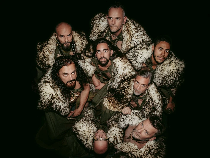 Corvus Corax 35 Jahre - Veranstaltung in der Leipzig Region Die sieben Männer der Corvus Corax in historisch anmutenden Kostümen mit Pelzumhängen schauen geschlossen und ernst in die Kamera, während sie im Kreis stehen. Das Bild ist dunkel gehalten und hebt die Gruppe in der Mitte hervor.