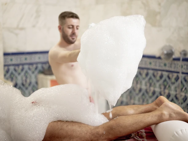 Mediterana Ein Mann führt eine Anwendung mit Schaum in der Sauna durch. Von einem zweiten Mann sind nackte Beine zu sehen.