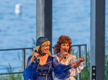 ABBA The Ladies am Biedermeierstrand - Veranstaltungen in der Leipzig Region Zwei Frauen als ABBA-Darstellerinnen singen und tanzen gemeinsam auf einer Open-Air-Bühne, beide tragen glitzernde, bunte Bühnenkostüme und halten jeweils ein Mikrofon in der Hand.