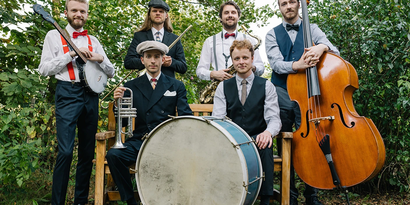 Rootbirds.jpg Sechs Musiker in Vintage-Kleidung mit Banjo, Trompete, Saxofon, Bass und großer Trommel im Freien