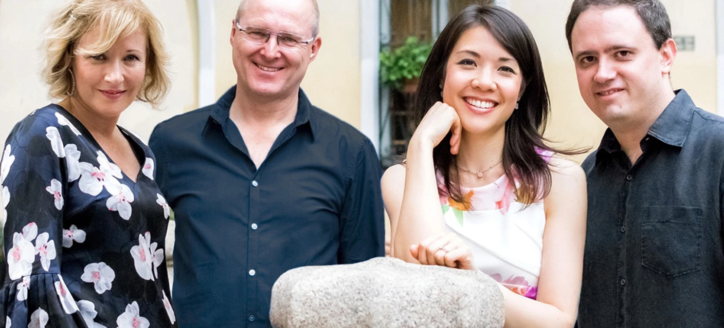 Swinging Ladies +2 Vier Erwachsene stehen eng beisammen und lächeln vor einem hellen Gebäude.