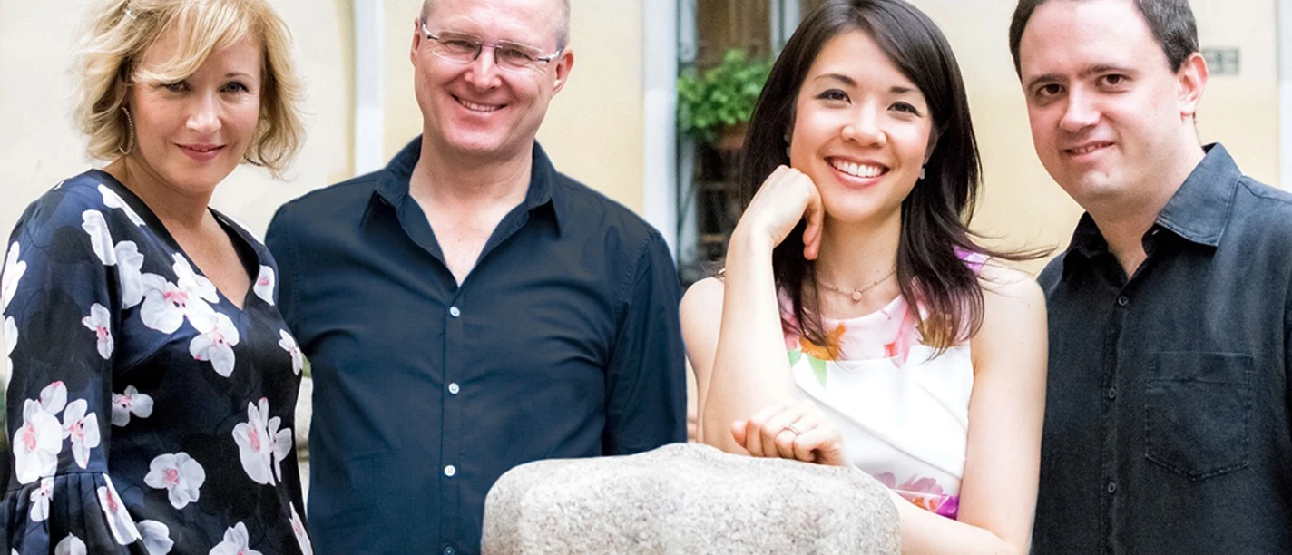 Swinging Ladies +2 Vier Erwachsene stehen eng beisammen und lächeln vor einem hellen Gebäude.