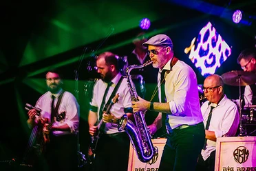 Big Brass Machine Mehrere Musiker spielen Saxofon, Gitarre und Schlagzeug auf einer bunt beleuchteten Bühne.