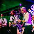 Big Brass Machine Mehrere Musiker spielen Saxofon, Gitarre und Schlagzeug auf einer bunt beleuchteten Bühne.