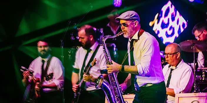 Big Brass Machine Mehrere Musiker spielen Saxofon, Gitarre und Schlagzeug auf einer bunt beleuchteten Bühne.