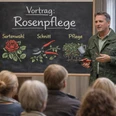 Rosenpflege Mann erklärt vor Tafel zu Rosenpflege einer sitzenden Gruppe mit Werkzeug und Rosenstock