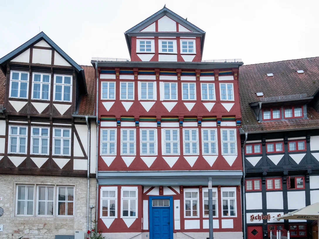 Schlossplatz18.jpg Fachwerkhaus mit roter und weißer Fassade, blauen Türrahmen und zahlreichen Fenstern am Schlossplatz.