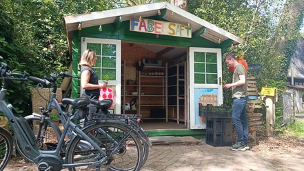 Hofladen auf dem Fabelhof Kleine Holzhütte mit Aufschrift „FABELSAFT“, zwei Personen davor mit Fahrrädern und Kisten.