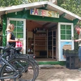 Hofladen auf dem Fabelhof Kleine Holzhütte mit Aufschrift „FABELSAFT“, zwei Personen davor mit Fahrrädern und Kisten.