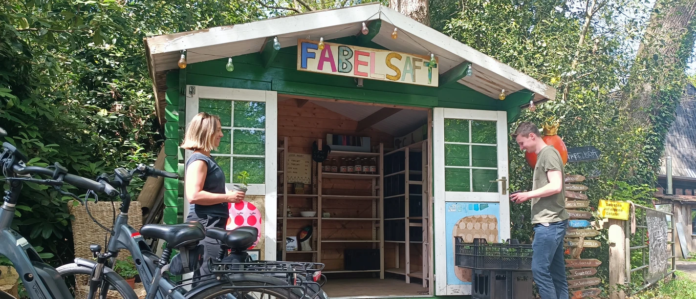 Hofladen auf dem Fabelhof Kleine Holzhütte mit Aufschrift „FABELSAFT“, zwei Personen davor mit Fahrrädern und Kisten.