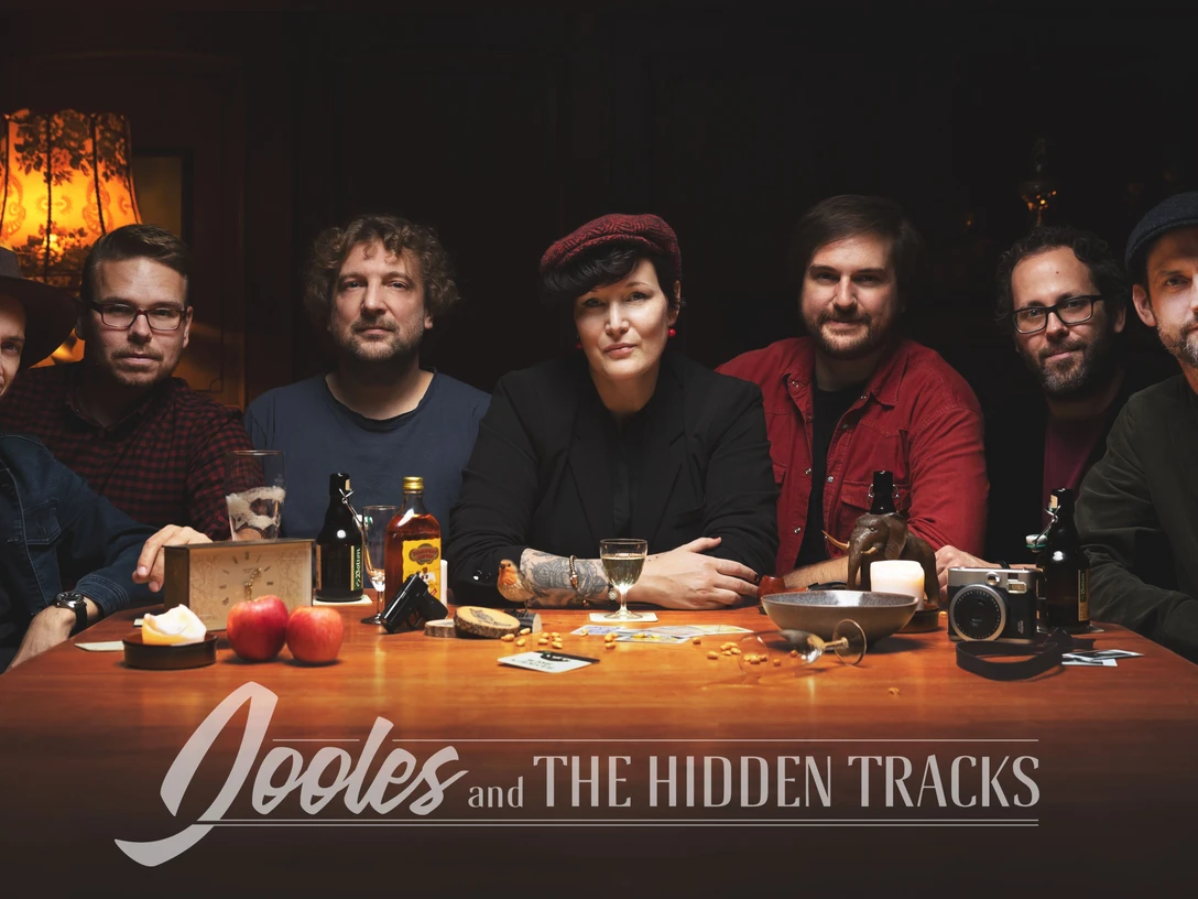 Sieben Bandmitglieder sitzen nebeneinander an einem Tisch mit Getränken. Hinter ihnen ein dunkler Raum mit Kaminfeuer.