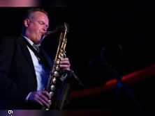 Saxophonist im dunklen Anzug spielt beleuchtet auf einer Bühne im Club