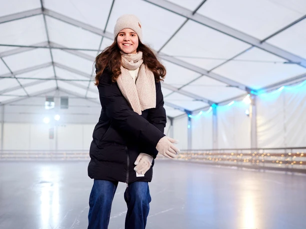 Frau in Winterkleidung lächelt beim Schlittschuhlaufen in einer überdachten Eisbahn.