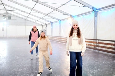 Drei Personen lächeln beim Schlittschuhlaufen in einer überdachten Eisbahn.